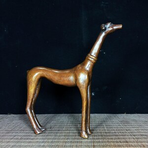 Könnte beinhalten: Eine braune Holzskulptur eines schlanken Hundes mit langem Hals und Beinen. Der Hund steht mit erhobenem Kopf. Die Skulptur hat eine glatte, polierte Oberfläche und steht vor einem dunklen Hintergrund.
