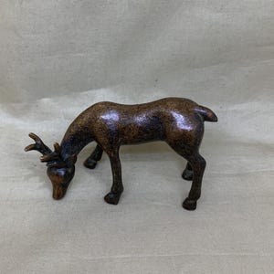 May include: A bronze-coloured deer figurine in a grazing pose. The deer has antlers and is bent over, appearing to be eating. The figurine is detailed with a textured surface and is set against a neutral background.