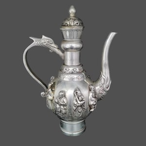 Op de afbeelding: Een zilveren theepot met een drakenvormige handvat en ingewikkelde houtsnijwerk van figuren. De theepot heeft een deksel met een knop en een rond lichaam.