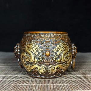 Może przedstawiać: Brązowa, okrągła miska dekoracyjna ze skomplikowanymi wzorami smoków i fal. Misa posiada dwie figury smoka i uchwyty pierścieniowe po każdej stronie. Projekt jest szczegółowy z ciemnobrązowym tłem.