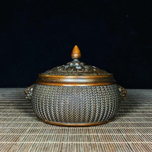 Może przedstawiać: Brązowy pojemnik z pokrywką o wzorze plecionego kosza. Pokrywka posiada skomplikowane rzeźbienia i mały, spiczasty szczyt. Pojemnik ma dwa uchwyty w kształcie głowy lwa po obu stronach. Element jest eksponowany na bambusowej macie.