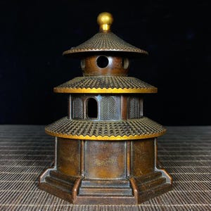 Może przedstawiać: Brązowy palnik do kadzidła w stylu pagody o wielopoziomowej konstrukcji. Konstrukcja posiada szereg dachów ze skomplikowanymi detalami i złotym zwieńczeniem. Element ma wygląd postarzałego, sugerujący wiek, i jest umieszczony na ciemnym tle.