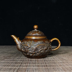 May include: Bronze-coloured teapot with detailed dragon relief carvings. The teapot has a curved spout, a rounded body, and a small handle. The lid is decorated with a small knob. The dragon design covers the body of the teapot.