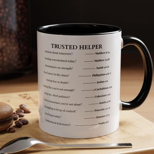 Pode incluir: Caneca de cerâmica branca com interior e alça pretos. A caneca apresenta o texto "TRUSTED HELPER" e uma lista de perguntas com versículos bíblicos correspondentes. Uma colher e um pastelaria estão em uma superfície de madeira.