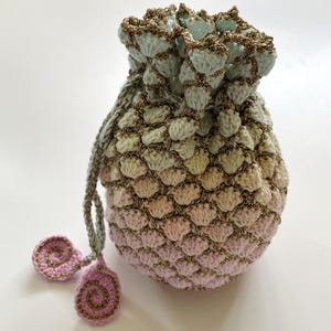 May include: A crocheted drawstring bag with a light blue, green, and pink ombre design. The bag has a gold glitter thread woven throughout the fabric. The bag has two small crocheted tassels.