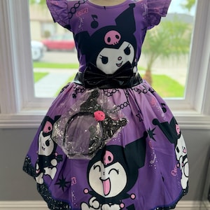 May include: A purple dress featuring a repeating print of the character Kuromi, musical notes, and stars. The dress has short ruffled sleeves, a black waistband with a bow, and a matching black headband in a plastic bag attached to the front.
