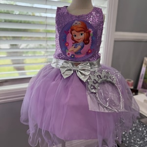 Pode incluir: Um vestido de princesa lavanda com um corpete de lantejoulas com um gráfico de personagem. O vestido tem um laço prateado e uma saia de tule em camadas. Uma tiara prateada está incluída.