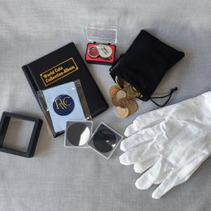 Puede incluir: Un conjunto de suministros para coleccionar monedas, que incluye un álbum negro con la etiqueta "World Coin Collection-Album", una bolsa de terciopelo negro llena de monedas, guantes blancos y soportes para monedas.
