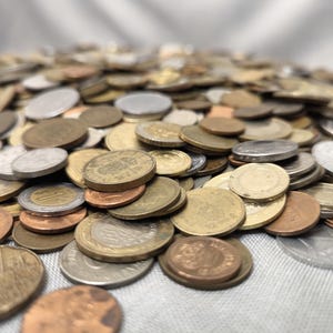 Può includere: Primo piano di un mucchio di monete di varie dimensioni, colori e forme. Le monete sono in argento, oro e rame e sono sparse su una superficie di colore chiaro.