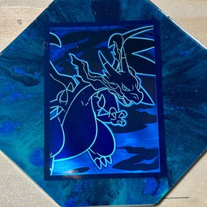 May include: A blue and black Pokemon card featuring a Charizard illustration. The card is set on a blue and teal resin coaster. The card has the text "©2023 Pokémon/Nintendo/Creatures/GAME FREAK."