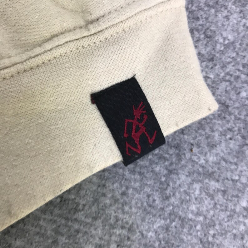 May include: Close-up of a cream-coloured fabric with a black tag. The tag features a red graphic of a person in motion. The fabric appears to be part of a garment, possibly a sweatshirt or jacket.