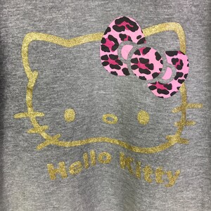 May include: Gray sweatshirt featuring a gold glitter outline of a cat face with a pink and black leopard print bow. The words "Hello Kitty" are written in gold glitter below the face.