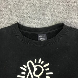 K&ouml;nnte beinhalten: Schwarzes Sweatshirt mit Rundhalsausschnitt, wei&szlig;em Grafikdesign und dem Text "K. Haring". Das Sweatshirt hat einen gerippten Ausschnitt und B&uuml;ndchen. Die Grafik ist eine stilisierte Figur mit Strahlenlinien.