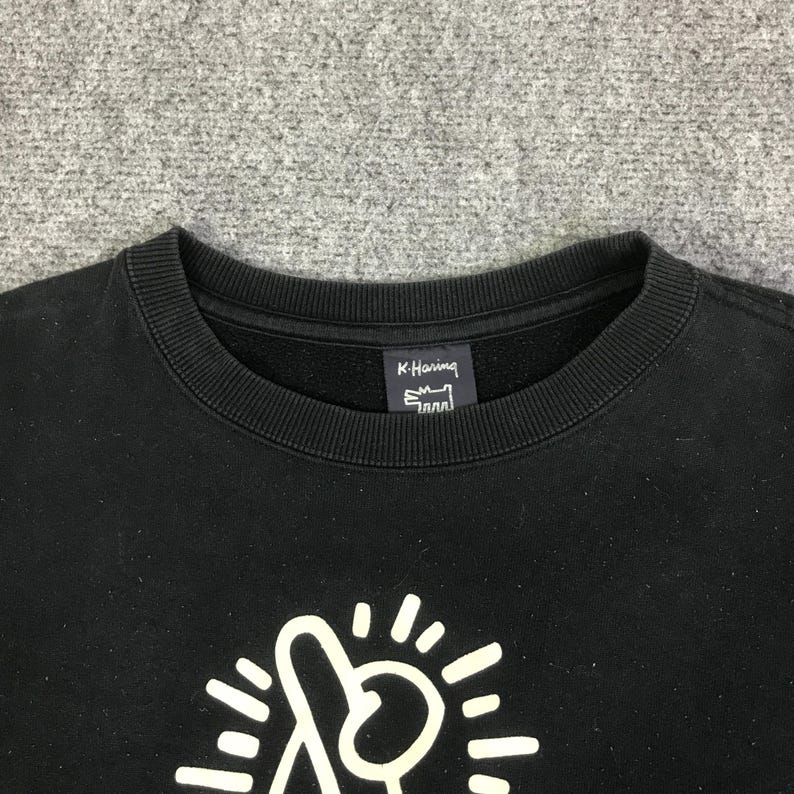 K&ouml;nnte beinhalten: Schwarzes Sweatshirt mit Rundhalsausschnitt, wei&szlig;em Grafikdesign und dem Text "K. Haring". Das Sweatshirt hat einen gerippten Ausschnitt und B&uuml;ndchen. Die Grafik ist eine stilisierte Figur mit Strahlenlinien.