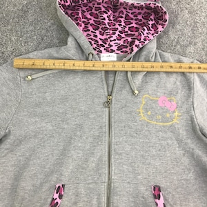 May include: A gray zip-up hoodie with a pink and black leopard print lining in the hood and pocket accents. The hoodie features a gold Hello Kitty graphic with a pink bow on the front.