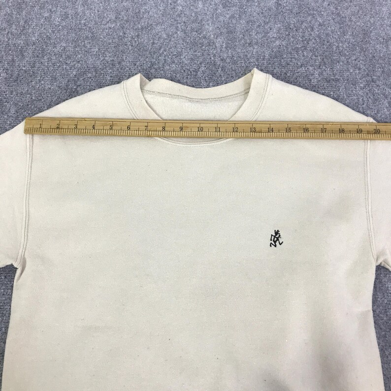 May include: Cream-coloured sweatshirt with a crew neck and long sleeves. A wooden ruler is placed across the top of the sweatshirt. A small black embroidered logo is on the left chest. The sweatshirt measures approximately 51 cm across.