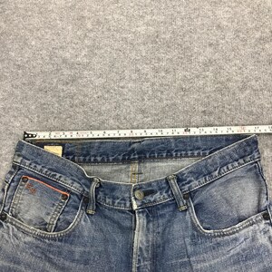 May include: Close-up of blue denim jeans with a button closure and front pockets. A measuring tape shows the waistline is approximately 17 inches across. The jeans have a faded wash and a small logo on the front pocket.