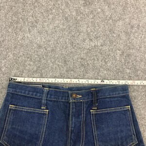 May include: Close-up of a pair of dark blue denim jeans. The jeans have a button closure, two front pockets, and yellow stitching. A measuring tape shows the waist is approximately 16 inches wide.