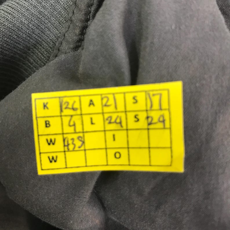 May include: A dark gray fabric garment featuring a yellow rectangular tag with a grid containing handwritten measurements. The grid displays values including K 26, A 21, S 17, B 4, L 24, S 24, and W 438, providing specific garment dimensions in inches.