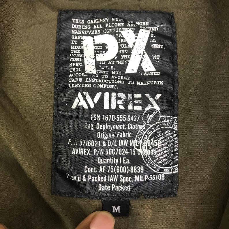 May include: A rectangular black fabric label sewn onto olive green material. The label features white text including the letters PX, the brand name AVIREX, and various military-style specifications such as FSN 1670-555-6437, P/N 57J6021, and a size M tag.