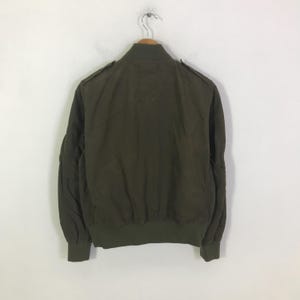 May include: An olive green bomber jacket featuring a ribbed collar, cuffs, and hem. The jacket includes shoulder epaulets and a full-length front zipper closure. The design is plain with long sleeves and a structured fit, presented on a hanger.