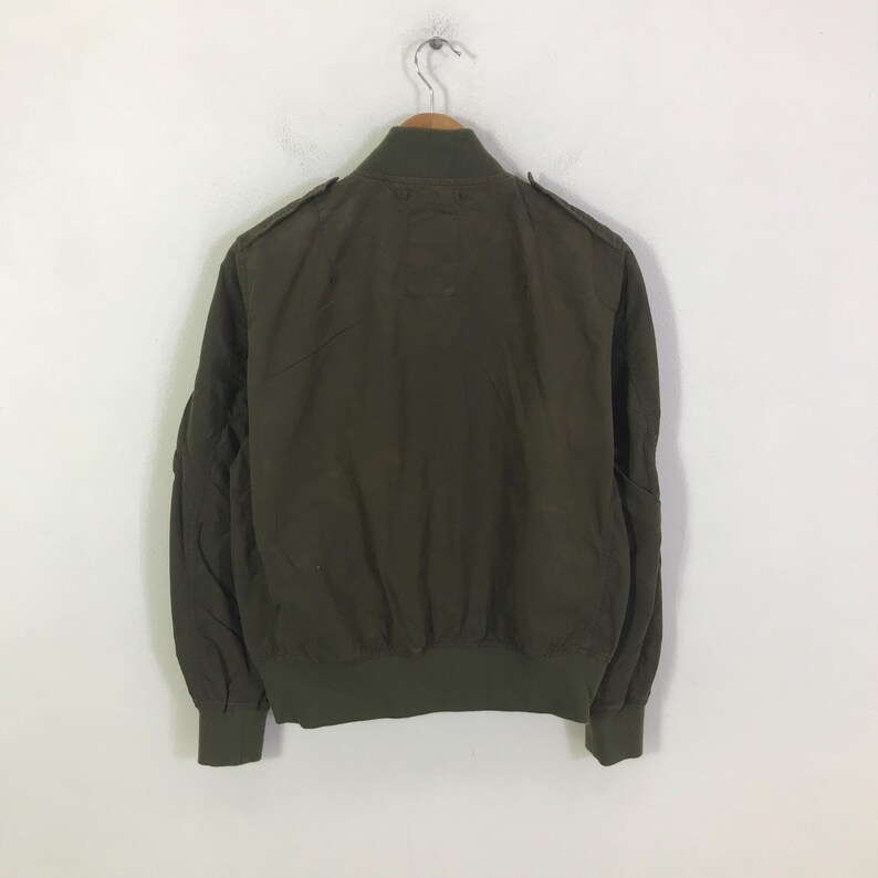 May include: An olive green bomber jacket featuring a ribbed collar, cuffs, and hem. The jacket includes shoulder epaulets and a full-length front zipper closure. The design is plain with long sleeves and a structured fit, presented on a hanger.