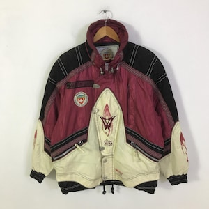 May include: A vintage ski jacket in burgundy, black, and cream. The jacket features a high collar, a Swiss emblem, and the word "Swiss" on the front. It has a zip closure and multiple pockets, with a geometric design.