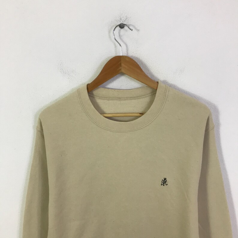 May include: A beige sweatshirt with a crew neck and long sleeves, suspended from a wooden hanger. A small, dark green embroidered logo is on the left chest. The sweatshirt is made of a soft material.
