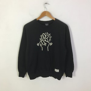 K&ouml;nnte beinhalten: Schwarzes Sweatshirt mit Rundhalsausschnitt und wei&szlig;er Grafik einer Hand, die ein gekreuztes Fingerzeichen macht. Das Sweatshirt h&auml;ngt an einem Holzkleiderb&uuml;gel. Ein kleines wei&szlig;es Etikett befindet sich auf der unteren rechten Seite.
