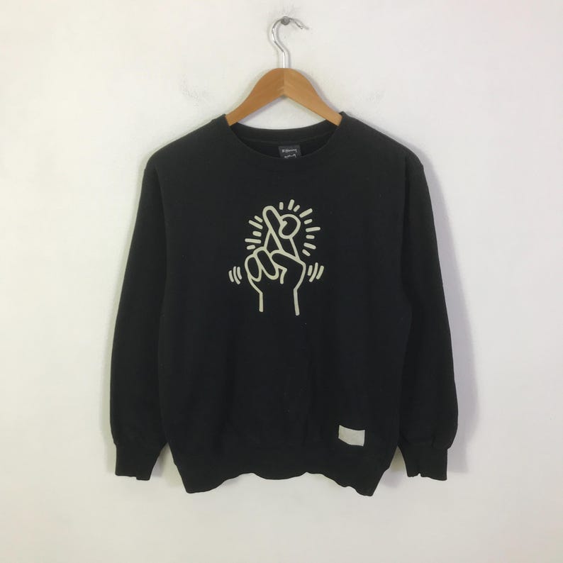 K&ouml;nnte beinhalten: Schwarzes Sweatshirt mit Rundhalsausschnitt und wei&szlig;er Grafik einer Hand, die ein gekreuztes Fingerzeichen macht. Das Sweatshirt h&auml;ngt an einem Holzkleiderb&uuml;gel. Ein kleines wei&szlig;es Etikett befindet sich auf der unteren rechten Seite.
