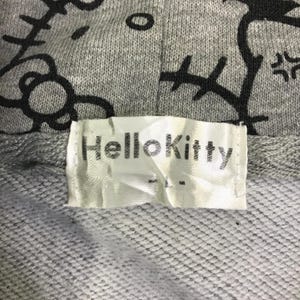 May include: Close-up of a gray hooded sweatshirt with a black Hello Kitty design. The label reads "Hello Kitty" in black text on a white background. The fabric is a textured knit.