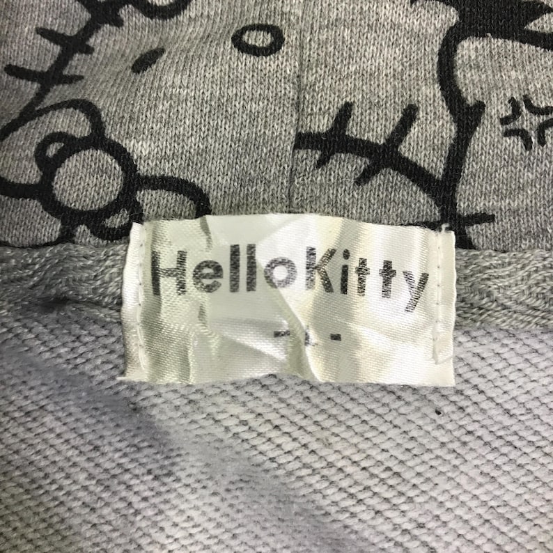 May include: Close-up of a gray hooded sweatshirt with a black Hello Kitty design. The label reads "Hello Kitty" in black text on a white background. The fabric is a textured knit.