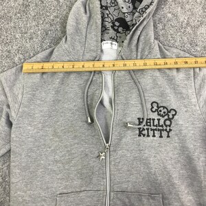 May include: A gray zip-up hoodie with a Hello Kitty skull design. The hoodie has a star-shaped zipper pull and a patterned hood lining. The ruler shows a length of approximately 17 inches.