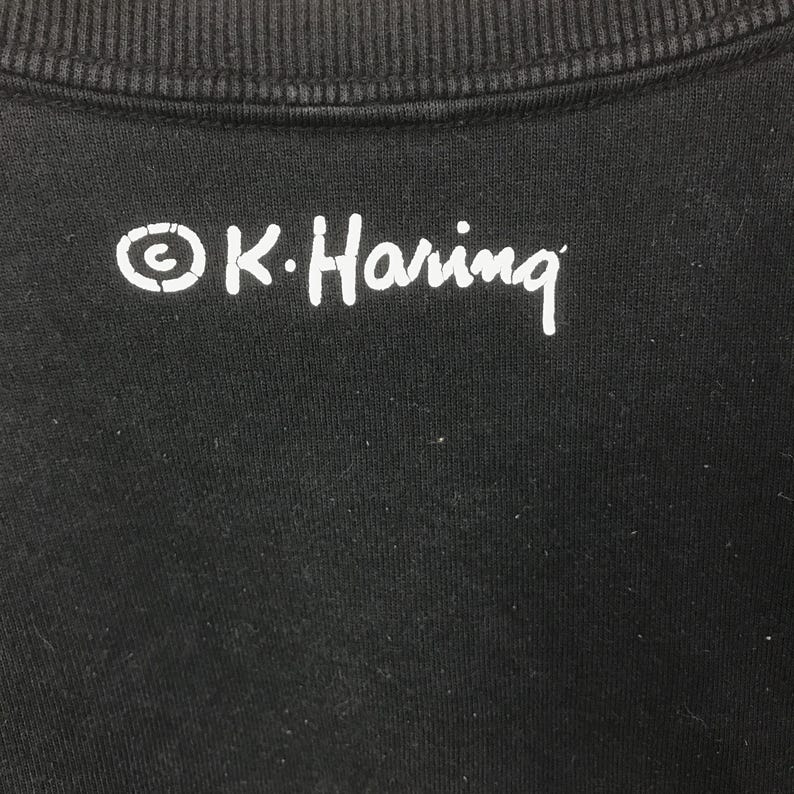 K&ouml;nnte beinhalten: Schwarzer Stoff mit dem wei&szlig;en Text "&copy; K. Haring" aufgedruckt. Der Text ist in handschriftlichem Stil gehalten. Der Stoff scheint ein Sweatshirt oder ein &auml;hnliches Kleidungsst&uuml;ck zu sein.