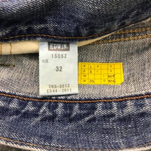 May include: Close-up of a pair of blue denim jeans with a white tag. The tag reads "EDWIN 1505Z" and "SIZE 32". A yellow tag with measurements is also visible. The jeans have a classic denim texture.