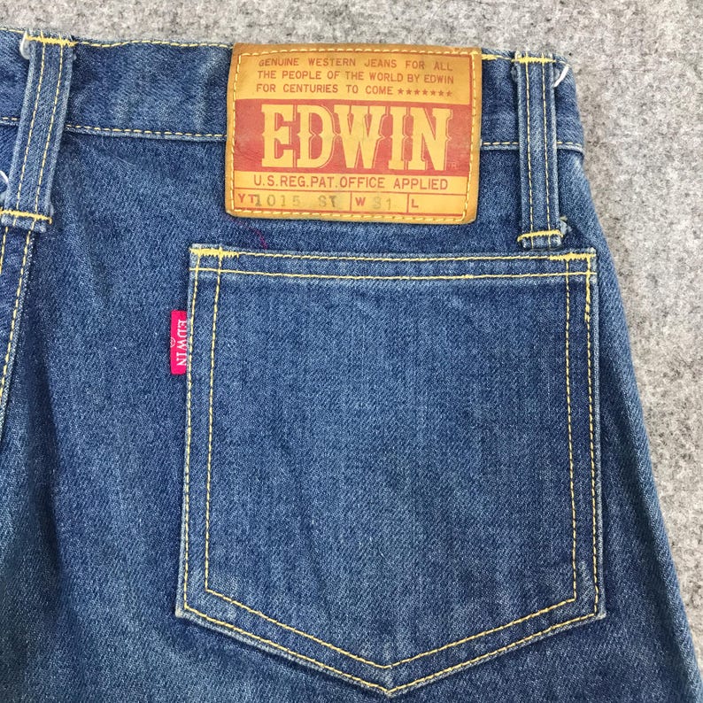 May include: Close-up of blue denim jeans with yellow stitching. The back pocket features a red Edwin tag. A large, rectangular leather patch with the Edwin logo and text is visible. The jeans are labeled "YT1015 ST W31 L".