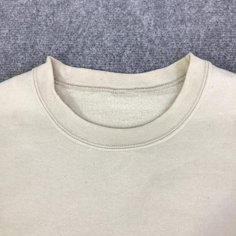 May include: A close-up shot of a light beige crew neck sweatshirt. The collar and neckline are visible, with a ribbed texture. The fabric appears soft and slightly textured, in a neutral tone. The background is a solid grey.