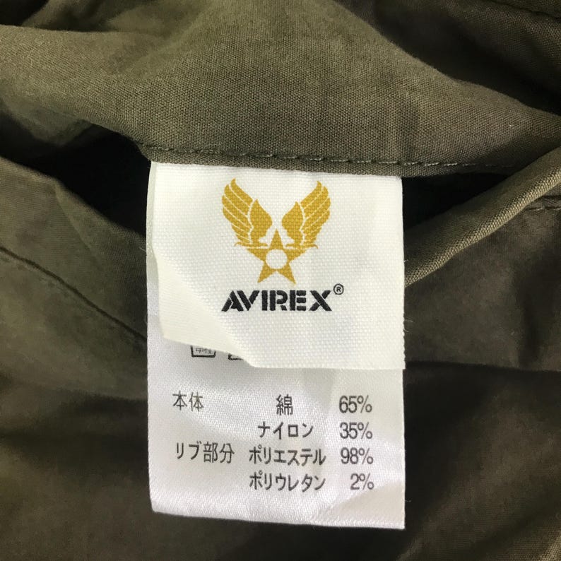 May include: A white fabric clothing tag sewn into an olive green garment. The tag features a yellow winged star logo above the text AVIREX. Below, Japanese text lists material composition as 65% cotton, 35% nylon, 98% polyester, and 2% polyurethane.