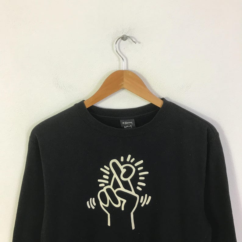 K&ouml;nnte beinhalten: Schwarzes Sweatshirt mit Rundhalsausschnitt und wei&szlig;er Grafik einer Hand, die ein Friedenszeichen zeigt. Das minimalistische Design zeigt strahlende Linien. Das Sweatshirt h&auml;ngt an einem Holzkleiderb&uuml;gel.