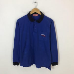 May include: A long-sleeved blue polo shirt with a black collar and cuffs. The shirt has a small pocket on the chest with red and white text. The collar has the text "TRD" in a red rectangle.