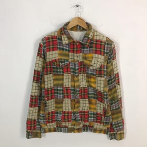Könnte beinhalten: Patchwork-Jacke mit verschiedenen Karomustern in Rot, Gelb, Grün und Beige. Die Jacke hat eine Knopfleiste, zwei Brusttaschen und lange Ärmel. Sie hängt an einem Holzkleiderbügel.