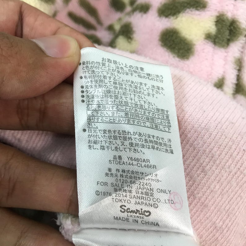 Puede incluir: Una etiqueta de tela rosa con texto japon&eacute;s y el logotipo de Sanrio. La etiqueta incluye instrucciones de cuidado e informaci&oacute;n del producto. El texto es negro y el logotipo es blanco.