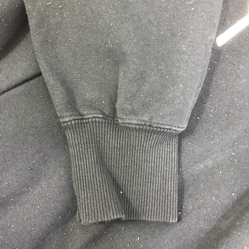 K&ouml;nnte beinhalten: Nahaufnahme eines dunkelgrauen Sweatshirt-&Auml;rmels. Das gerippte B&uuml;ndchen ist sichtbar, mit vertikalen Linien. Der Stoff hat ein leicht abgenutztes Aussehen mit kleinen wei&szlig;en Flocken.