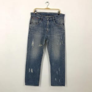 May include: Light blue denim jeans with distressed details, including rips and fading. The jeans are hanging on a gold-colored hanger. The jeans have a classic five-pocket design.