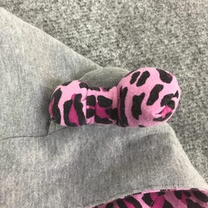 May include: A pink bow with a black leopard print pattern is attached to a gray fabric. The bow is made of a soft material and has a rounded shape. The fabric appears to be a sweatshirt material.