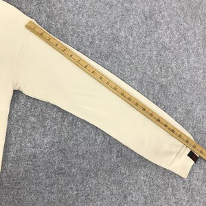 May include: Cream-coloured long-sleeved sweatshirt with a ruler across the sleeve. The sleeve has a red and navy blue stripe detail at the cuff. The sweatshirt is on a grey surface.