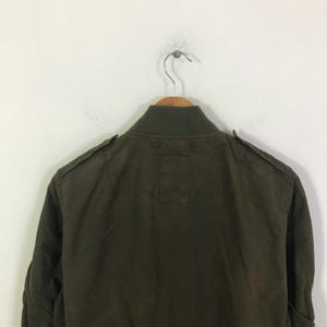 May include: An olive green bomber jacket featuring a ribbed collar, shoulder epaulets, and a plain back panel. The jacket is displayed on a wooden hanger, showcasing the solid color fabric and the structured design of the upper back and shoulders.