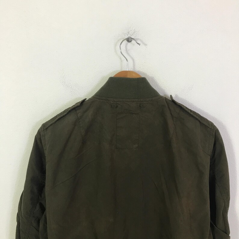 May include: An olive green bomber jacket featuring a ribbed collar, shoulder epaulets, and a plain back panel. The jacket is displayed on a wooden hanger, showcasing the solid color fabric and the structured design of the upper back and shoulders.