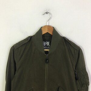 May include: An olive green bomber jacket featuring a ribbed collar, a full-length front zipper, and epaulets on the shoulders. The interior label displays the text PX and AVIREX. A zippered utility pocket is visible on the upper left sleeve.