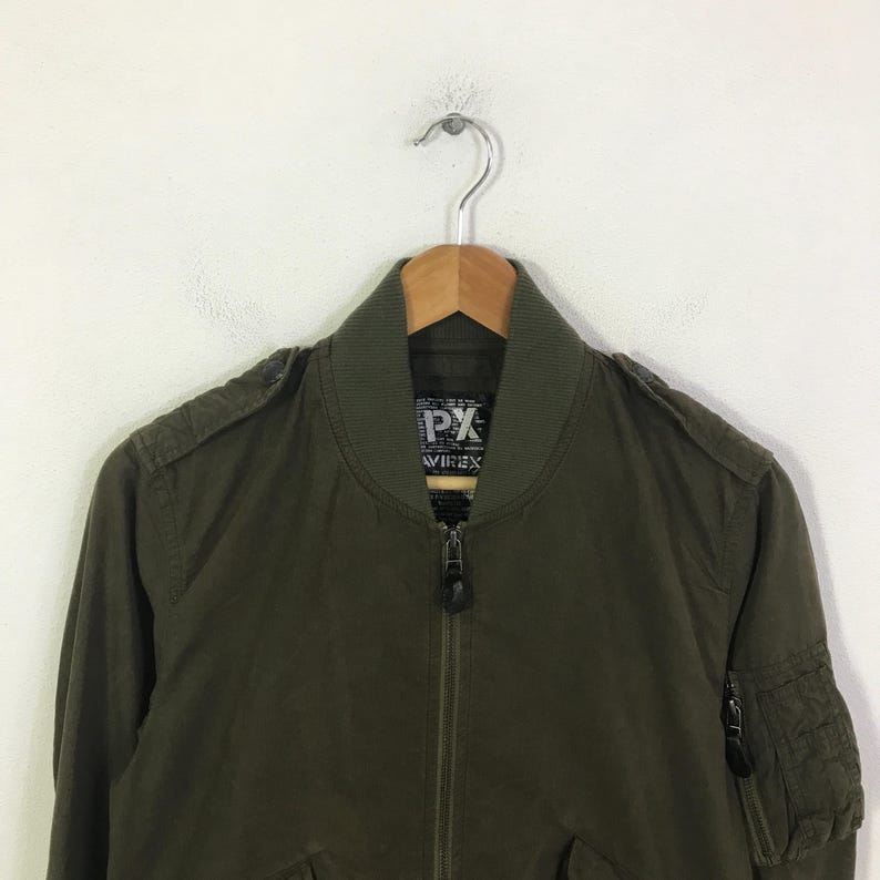 May include: An olive green bomber jacket featuring a ribbed collar, a full-length front zipper, and epaulets on the shoulders. The interior label displays the text PX and AVIREX. A zippered utility pocket is visible on the upper left sleeve.